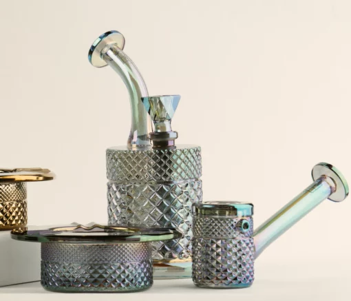 Three iridescent glass smoking accessories—a bong, a pipe, and the Twenties Collection Jar | Mermaid—are arranged on a light surface, each displaying a mermaid-inspired diamond-cut pattern and metallic sheen.