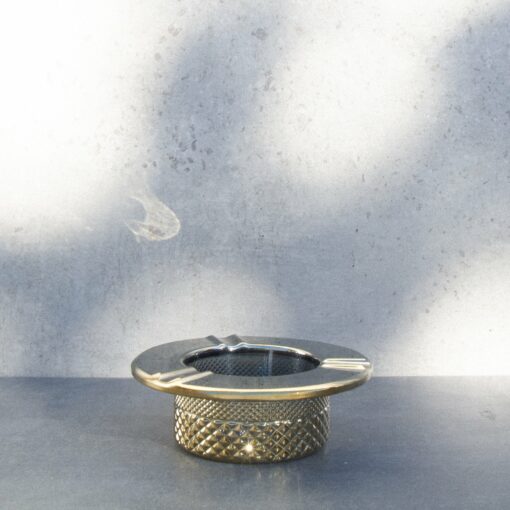 The Twenties Collection Ashtray | Gold, featuring a metallic textured finish, rests on a dark surface before a light grey concrete wall with soft shadows.
