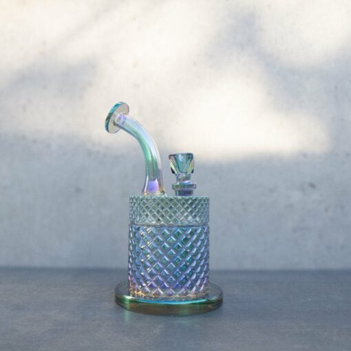 The Twenties Collection Water Pipe | Mermaid, a small iridescent glass piece with a diamond-patterned base, sits on a gray surface against a light, blurred background.