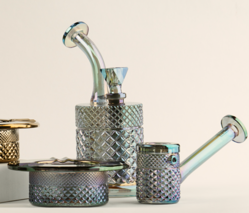 Two iridescent glass pipes from the Twenties Collection, one angled and one straight, are displayed with a Mermaid Ashtray from the same collection and a metal grinder against a light background. The pipes feature textured, diamond-patterned surfaces.