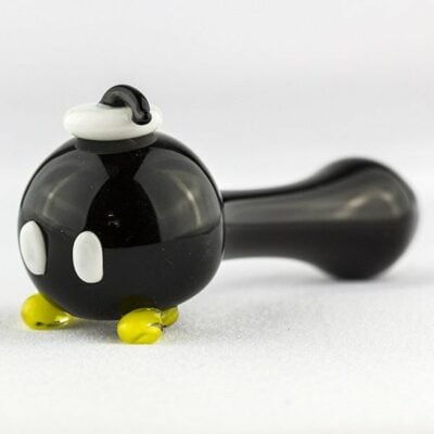 The Empire Glassworks Bomber Spoon Pipe is a glass pipe designed to look like a black cartoon bomb with white eyes, yellow feet, and a fuse cap, inspired by the Bob-omb character from video games.