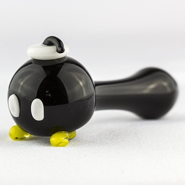 The Empire Glassworks Bomber Spoon Pipe is a glass pipe designed to look like a black cartoon bomb with white eyes, yellow feet, and a fuse cap, inspired by the Bob-omb character from video games.