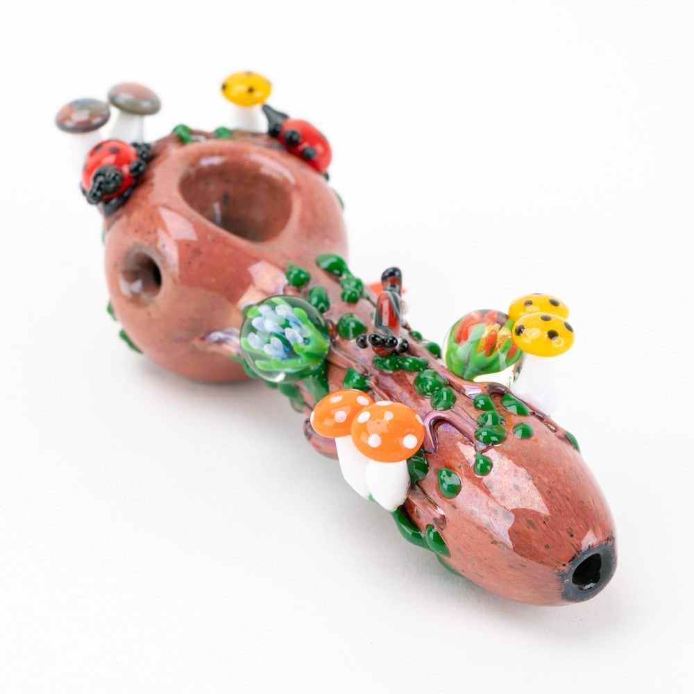 The Empire Glassworks Bug's Life Small Spoon Pipe is a decorative hand pipe designed to resemble a log, featuring colorful glass mushrooms, ladybugs, and green vines for a whimsical atmosphere.