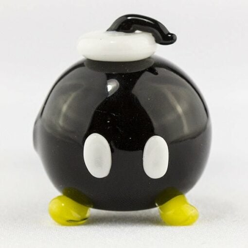 The Empire Glassworks Bomber Spoon Pipe is a small, round, black pipe with white oval eyes, yellow feet, a white fuse holder, and a curled black fuse on top, displayed against a plain white background.