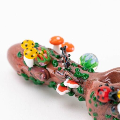 A close-up of the Empire Glassworks Bug's Life Small Spoon Pipe, crafted to resemble a log with vibrant glass mushrooms, leaves, and a ladybug, displayed on a white background.