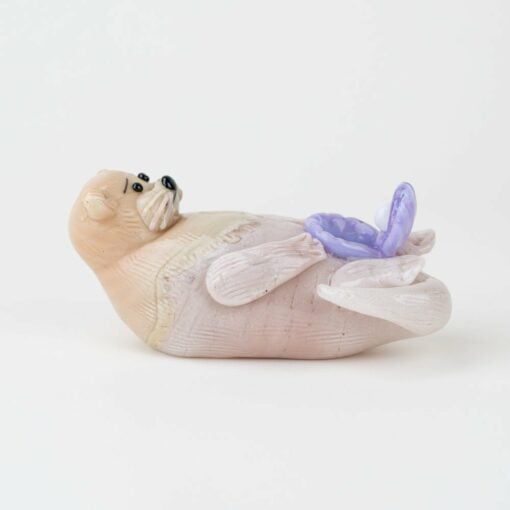 The Empire Glassworks Otter Dry Pipe features a light brown and beige glass otter lying on its back, holding a purple seashell with a white pearl inside, all set against a plain white background.