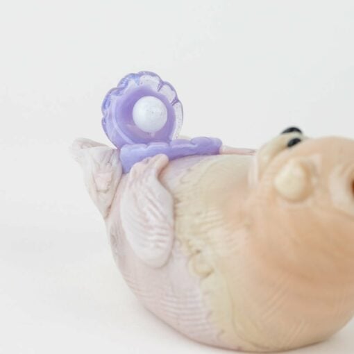 The Empire Glassworks Otter Dry Pipe is a small, light brown pipe shaped like an otter lying on its back and holding a purple seashell with a white pearl inside, displayed against a plain white background.