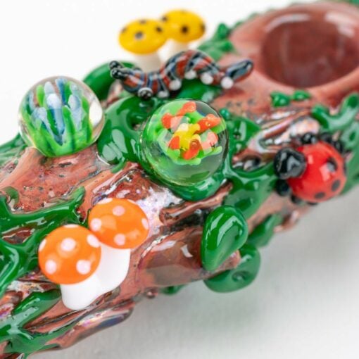 The Empire Glassworks Mossy Log Dry Pipe features a forest log design adorned with colorful glass mushrooms, a ladybug, a caterpillar, and decorative marbles mimicking plants and insects.