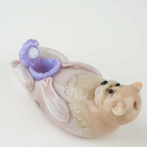 The Empire Glassworks Otter Dry Pipe features a pale, reclining ceramic otter holding a purple clam shell with a white pearl, set on a plain white background—ideal for fans of Empire Glassworks artistry.