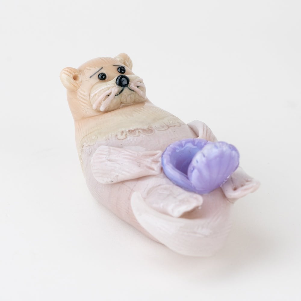 A small, light-colored Otter Dry Pipe by Empire Glassworks lies on its back holding a purple seashell on its belly, set against a simple white background.