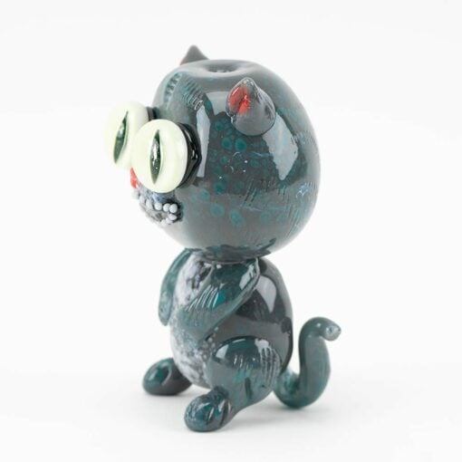 The Empire Glassworks Galacticat Dry Pipe is a small, glossy dark green glass pipe shaped like a cartoon cat with oversized light green eyes, a red ear spot, upright posture, and curled tail, displayed against a white background.