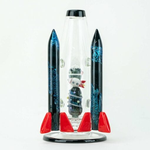 The Empire Glassworks Galactic Flagship Water Pipe sports a glass rocket design with black and blue glitter fins, red cone tips, and a cartoon bear inside standing upright against a white background.