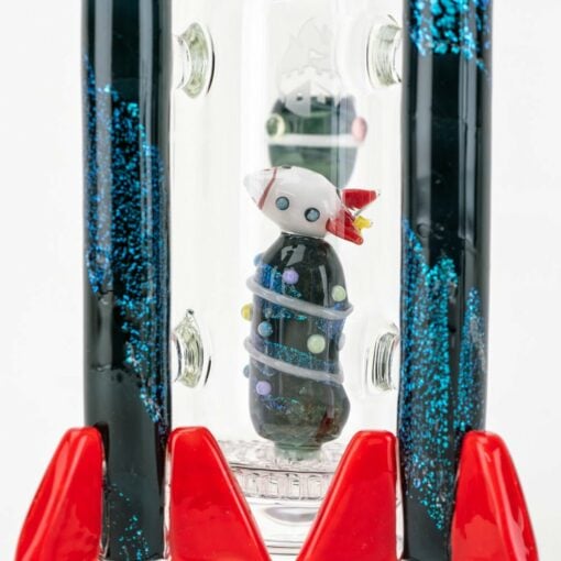 The Empire Glassworks Galactic Flagship Water Pipe boasts a whimsical rocket design with red fins, sparkling black sides accented by blue glitter, and a clear chamber containing a small wrapped figure with a dotted white head.