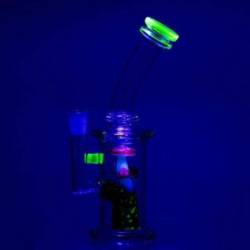 The Empire Glassworks Cosmic Mushrooms Mini Rig showcases neon green accents and an internal mushroom design that glows brightly under blue or blacklight, creating a vivid effect against dark backgrounds.