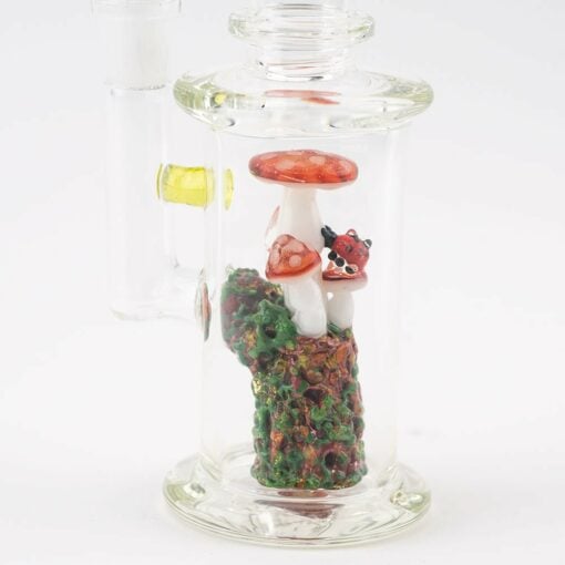 The Empire Glassworks Cosmic Mushrooms Mini Rig is a clear glass piece featuring red mushrooms, a small red ladybug, and a green and brown textured base, all showcased against a white background.