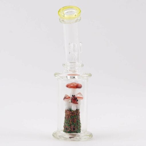 The Empire Glassworks Cosmic Mushrooms Mini Rig showcases a clear chamber with a Cosmic Mushrooms scene, a small white figure on a textured green base, and a yellow-tinted mouthpiece rim for added style.