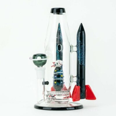 The Empire Glassworks Galactic Flagship Water Pipe is a glass bong shaped like a rocket ship, with red fins, a black nosecone, and a tiny astronaut inside. Space-themed details and glitter complete this stunning piece against a white background.