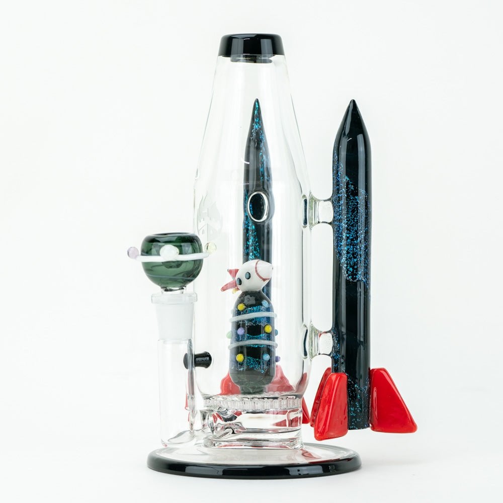 The Empire Glassworks Galactic Flagship Water Pipe is a glass bong shaped like a rocket ship, with red fins, a black nosecone, and a tiny astronaut inside. Space-themed details and glitter complete this stunning piece against a white background.
