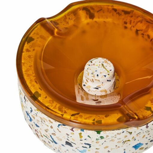 A close-up of the NWTN Home Symi Ashtray shows its round, speckled body with an amber transparent lid, central raised knob, and two spout-shaped indentations on the edge.