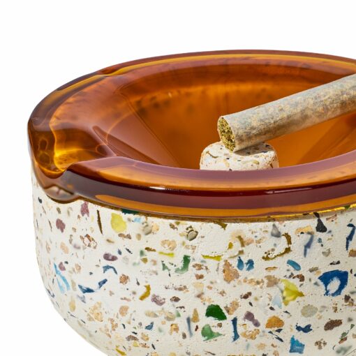 A close-up of the NWTN Home Symi Ashtray highlights its terrazzo pattern with colorful stone fragments in a white base and a translucent amber glass rim, featuring a groove that holds a rolled cigarette.