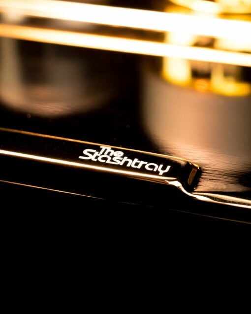 Close-up of a glossy black Stashtray with “The Stashtray” printed in white, reflecting warm yellow lighting—the perfect accessory for the 24k Stashtray Bundle.