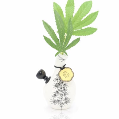 The Love Bud Vase Bong is a white ceramic vase shaped like a small bong with a black floral design, holding green leaves at its opening. A gold "with love" tag hangs from the neck, set against a minimalistic white background.