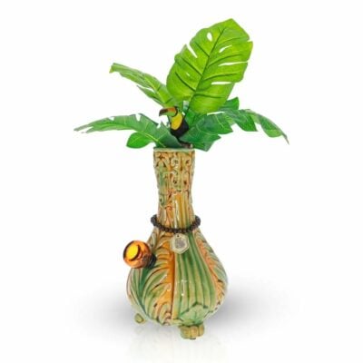 The TocaCabana Vase Bong is a decorative ceramic piece with green and brown leaf patterns, artificial tropical leaves, a small toucan figurine, and an orange charm hanging from a black cord around the neck.