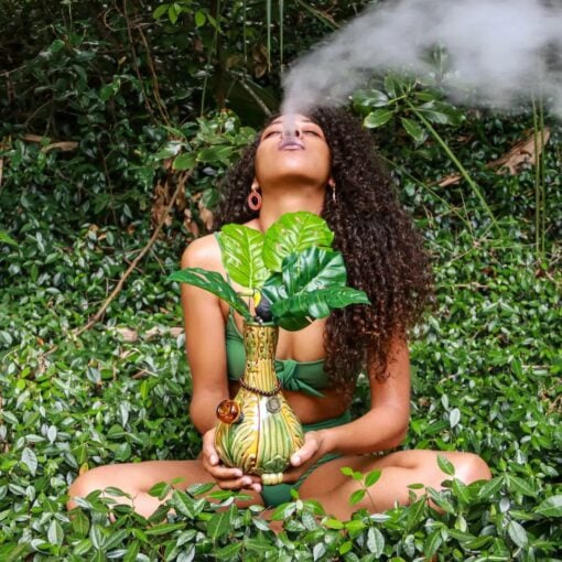 A woman in a green bikini sits cross-legged amid lush greenery, holding the TocaCabana Vase Bong—designed to resemble a plant—and exhales a thick cloud of smoke as she’s surrounded by dense foliage.