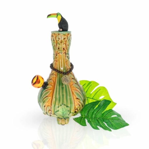 The TocaCabana Vase Bong features a green leaf pattern, a small toucan figurine on top, a beaded necklace with round pendant, and a yellow-orange bowl, all displayed with tropical leaves for perfect TocaCabana vibes.
