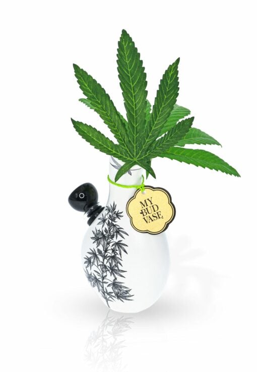 A small white Love Bud Vase Bong with a black bamboo design holds cannabis leaves. A gold MY BUD VASE tag hangs around its neck. The background is white and minimalistic.