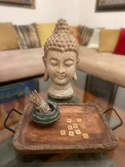 A Buddha head statue sits on a table with the Woodland Rolling Tray, which holds sage, a small bowl, and Scrabble tiles spelling MY BUD VASE. A beige sofa with colorful cushions appears in the background.