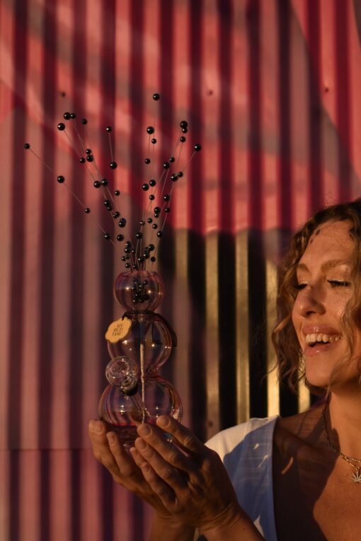 A woman with curly hair smiles and holds the Bubbles Vase Bong with black decorative sticks, standing in front of a corrugated wall where warm sunlight sparkles through bubbles, creating pink and black shadows.