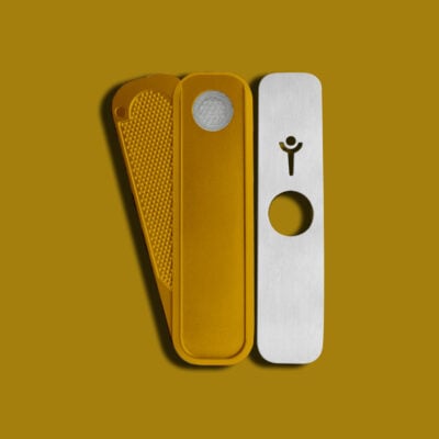 A mustard-yellow metal herb grinder, designed to pair with the Gold Mini Genius Pipe, has a textured grip, mesh screen, and a lid featuring a circular hole and small person icon—all on a mustard-yellow background.