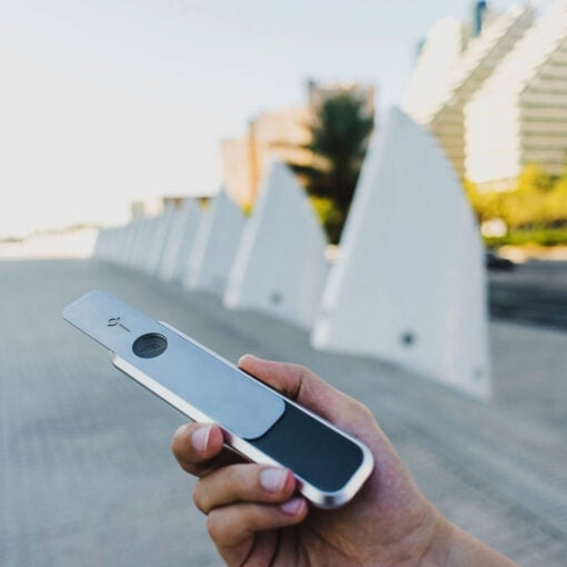A hand holds the High Expectations Genius Pipe outdoors, with geometric white structures and blurred buildings in the background—evoking a sense of smart, sophisticated living.