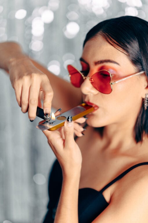 A woman with short dark hair and red-tinted heart-shaped sunglasses lights a Gold Mini Genius Pipe. She wears a black top, has black and white painted nails, and stands before a sparkly, blurred background.