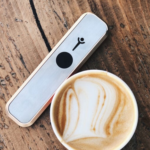 A High Expectations Genius Pipe, a sleek rectangular metal smoking pipe with a black dot and symbol, rests on a wooden table beside a latte topped with intricate milk froth art.