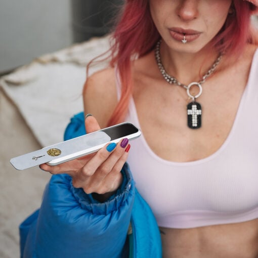 A person with pink hair and colorful nails holds a metal tray with ground cannabis and a High Expectations Genius Pipe, wearing a cross necklace, white top, and blue jacket draped over one shoulder.