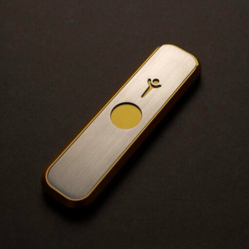A close-up of the Gold Mini Genius Pipe, a sleek rectangular metallic device with rounded edges, a yellow circular button, and a small abstract symbol on its brushed surface, set against a dark background.