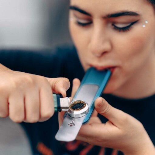 A person uses a lighter to ignite material in a Blue Mini Genius Pipe, holding it to their mouth and inhaling. They wear bold makeup with winged eyeliner and face jewels.