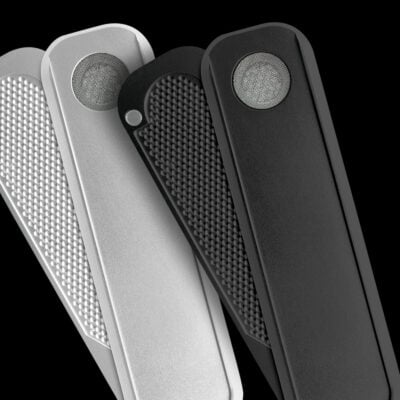 Three sleek metal objects, including the Black Mini Genius Pipe and Black Genius Pipe, feature textured and smooth surfaces with a circular mesh detail at the top. The pieces are silver, gray, and black against a black background.