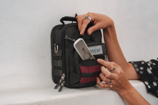 A person with several rings and patterned sleeves places a portable medical device labeled “green us” into the front pocket of a small black and red backpack.