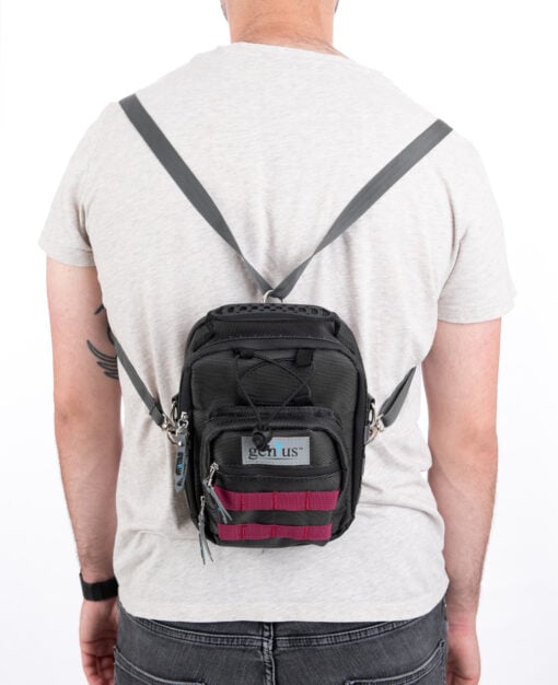 A person in a light gray t-shirt and jeans faces away from the camera, showcasing the Backpack—a small black bag featuring gray straps and red accents.