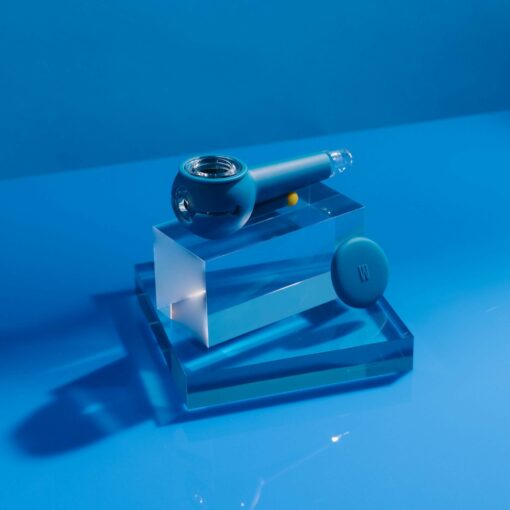 The Pipe - Midnight, a modern midnight blue glass pipe, is showcased on clear acrylic blocks with a round blue container against a bright blue background.