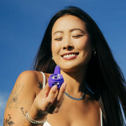 A woman with long dark hair and a tattoo on her arm smiles brightly outdoors, squinting under the blue sky as she holds The Pipe - Grape in her hand.