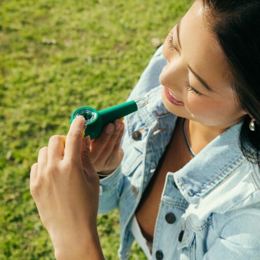 A woman wearing a denim jacket smiles as she lights The Pipe - Forest outdoors, holding the green pipe in one hand on a grassy area near the edge of a forest.