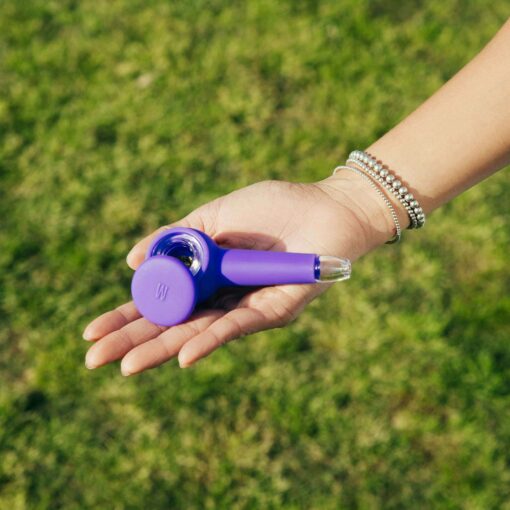 A hand with silver bracelets holds The Pipe - Grape, a small smoking pipe with a transparent tip, set against a blurred green grass background.