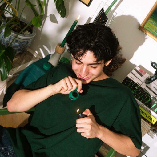 A young person in a green shirt sits indoors, smiling as they light The Pipe - Forest with a lighter. Plants, green candles, and stacked books are visible in the background.