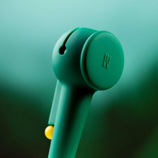 A close-up of The Pipe - Forest, a teal minimalist earbud with a circular end and subtle "W" logo. The softly blurred green-blue background evokes the tranquility of a forest.
