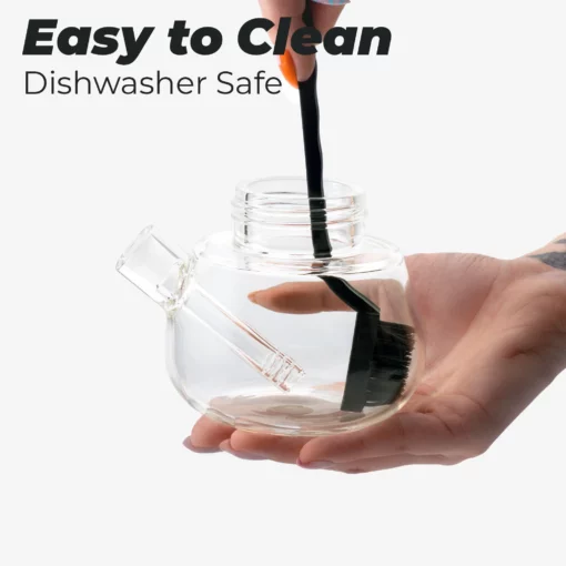 A person cleans the interior of The Bubbler - Grape, a clear glass teapot with a grape-inspired design, using a black brush. Text above reads "Easy to Clean Dishwasher Safe." The background is white.