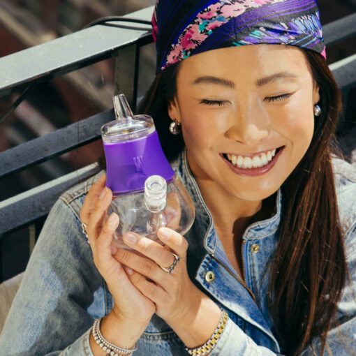 A smiling woman with long dark hair and a colorful headband holds The Bubbler - Grape, eyes closed, wearing a denim jacket and bracelets while standing outdoors by a metal railing.